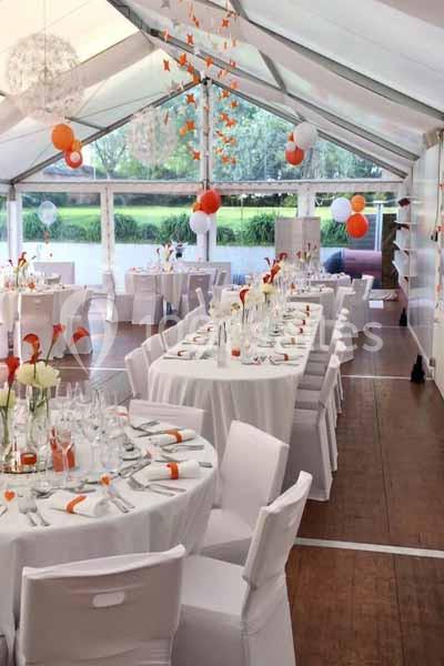 Salle de réception sous une tente avec tables rondes décorées de nappes blanches et accents orange.