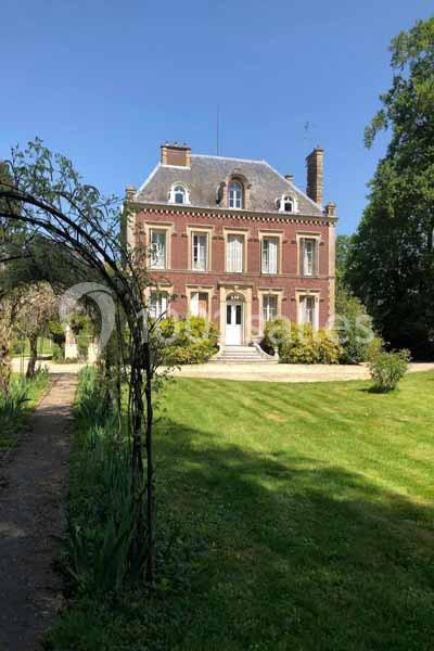 Maison en briques rouges à deux étages, entourée d'un jardin verdoyant avec une arche végétale au premier plan.