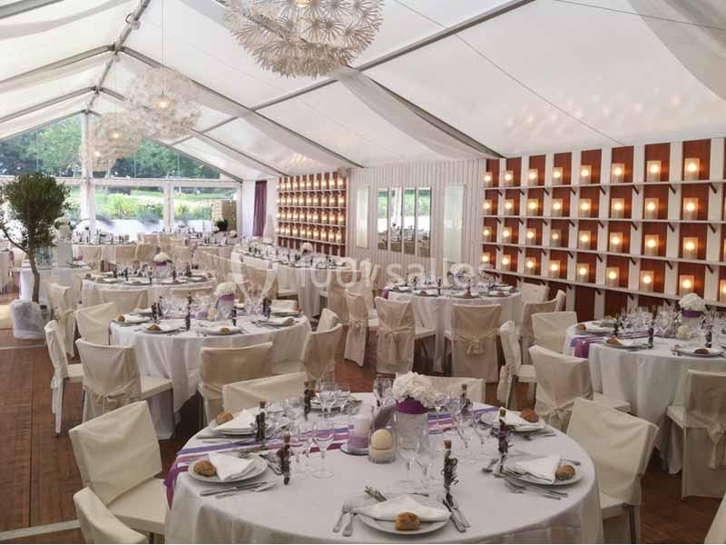Salle de réception sous une tente blanche, avec tables rondes dressées et décorations élégantes.