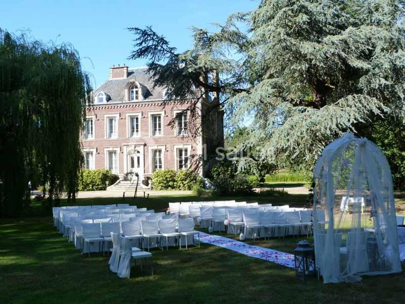 Chaises blanches disposées en extérieur devant un manoir en briques, entouré d'arbres, pour une cérémonie.