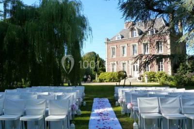 Glycine en fleurs grimpant sur une pergola, avec une maison en briques et des arbustes en arrière-plan.