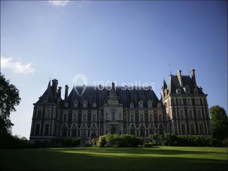 Façade d'un château historique en pierre, entouré de pelouses et d'arbres, sous un ciel bleu dégagé.