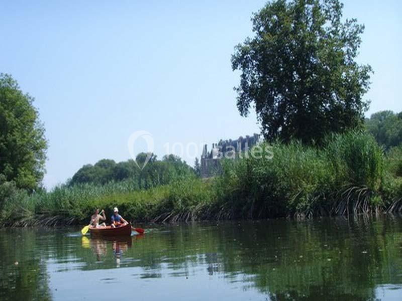Deux personnes pagayant dans une barque sur une rivière bordée de végétation, avec un château visible au loin.