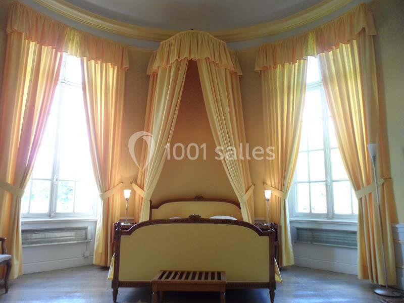 Lit à baldaquin en bois avec draperies jaunes, encadré par deux grandes fenêtres laissant entrer la lumière naturelle.