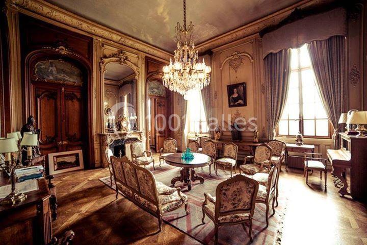 Salon ancien avec mobilier d'époque, parquet en bois, lustre en cristal et grandes fenêtres laissant entrer la lumière.