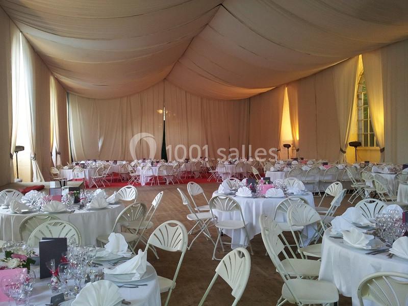 Salle de réception décorée avec des tables rondes dressées, nappes blanches et chaises pliantes, éclairage tamisé.