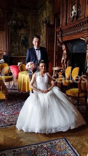 Un couple de mariés pose dans une salle ornée de meubles anciens et de tapisseries décoratives.