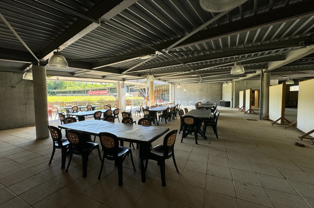 Salle couverte avec des tables et chaises en plastique noir, éclairée par des lampes suspendues et ouverte sur l'extérieur.