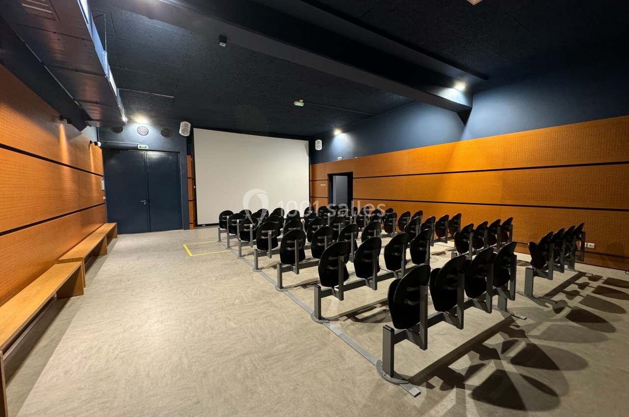 Salle de projection vide avec des rangées de sièges noirs, un écran blanc et des murs en bois clair.