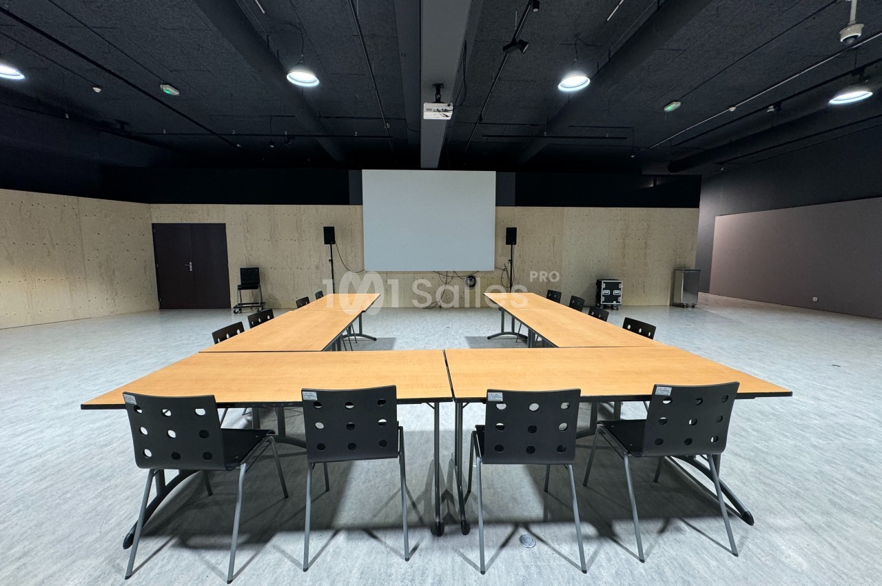 Salle de réunion avec des tables disposées en U, des chaises noires et un écran de projection au fond.