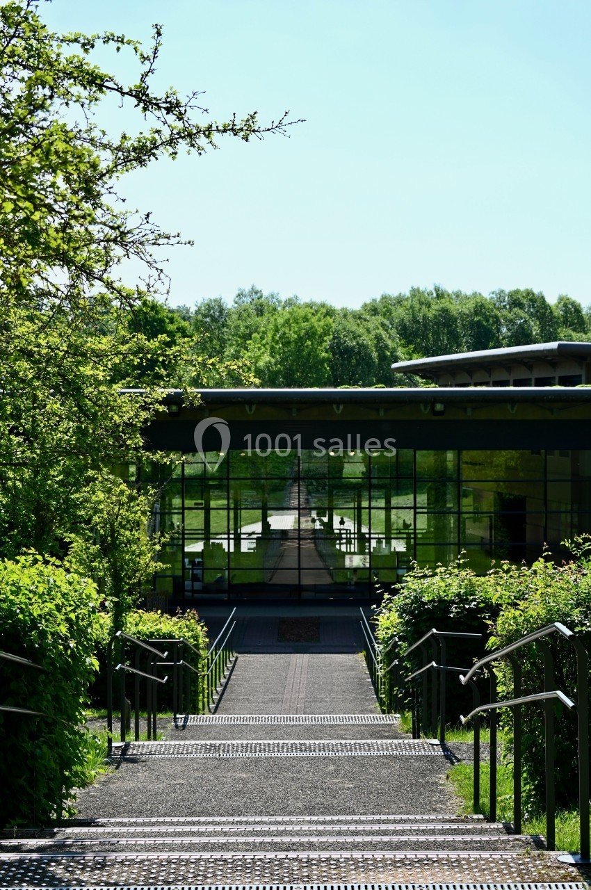 Escalier bordé de verdure menant à un bâtiment moderne avec une façade vitrée reflétant un paysage arboré.