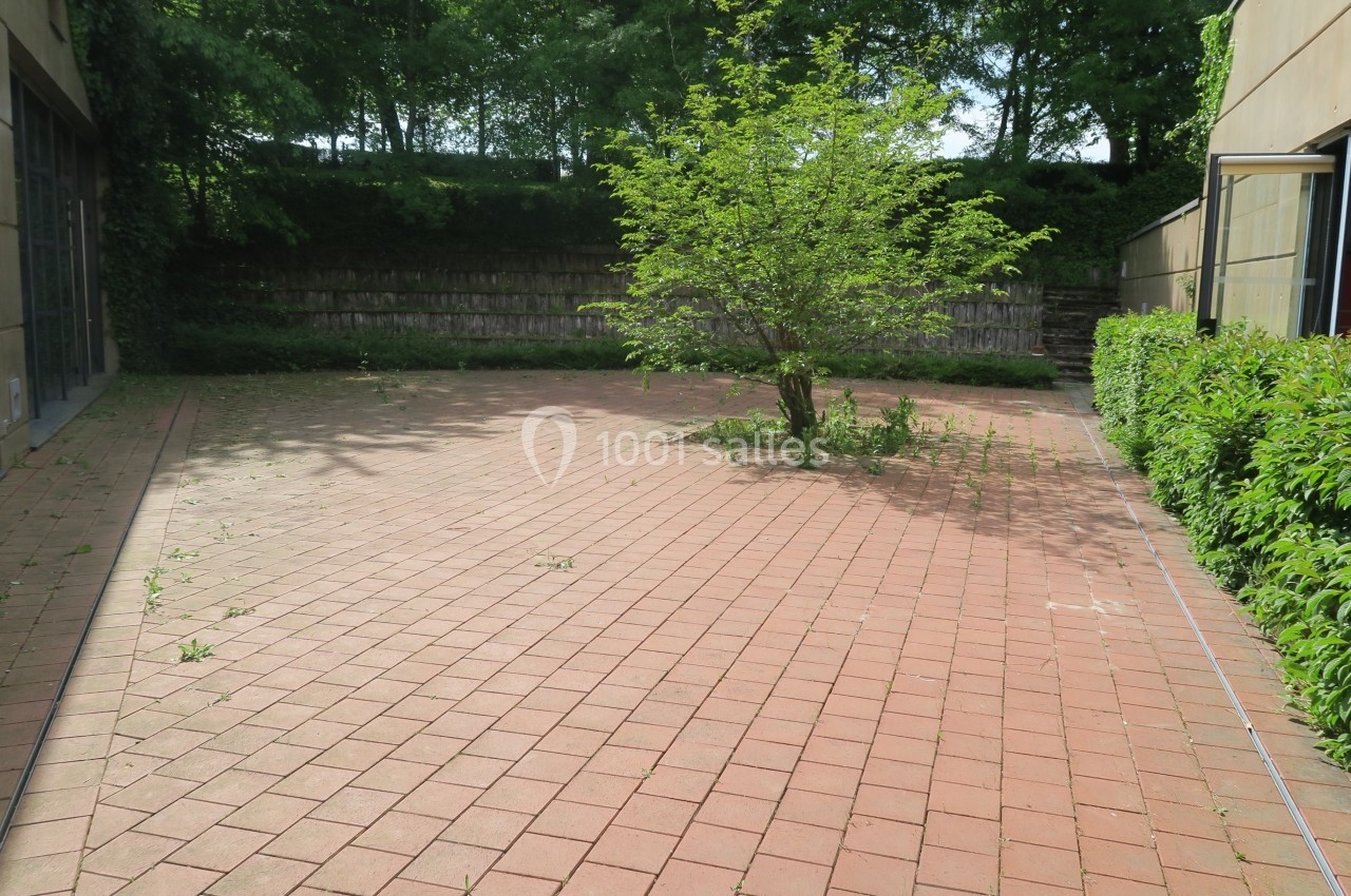 Cour pavée avec un petit arbre au centre, entourée de bâtiments et de haies verdoyantes sous un ciel dégagé.