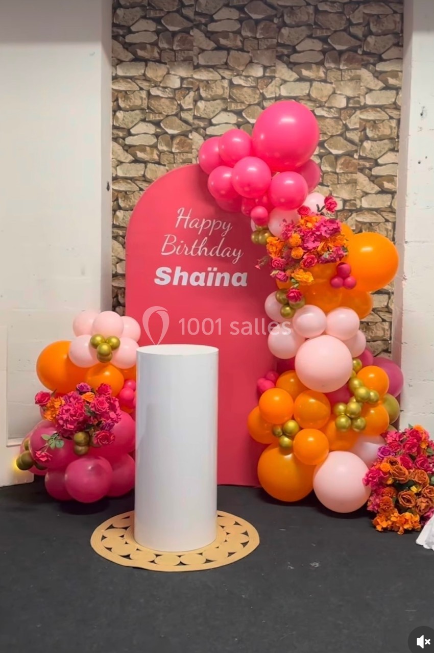 Décor d'anniversaire avec arche rose, ballons colorés, fleurs et un présentoir cylindrique blanc.