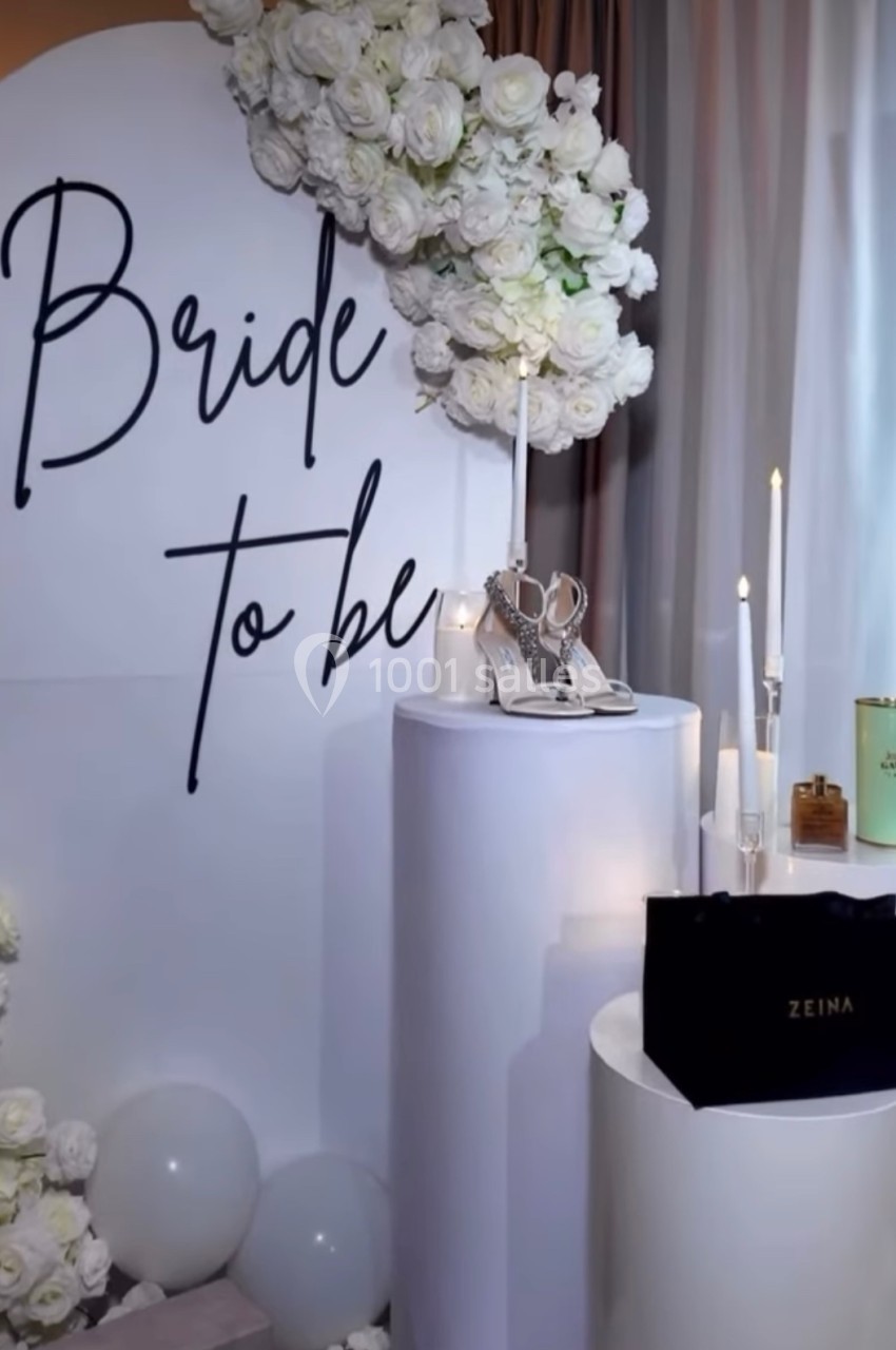 Décor élégant avec fleurs blanches, bougies, chaussures de mariée et sac noir, sur fond ’Bride to be’.