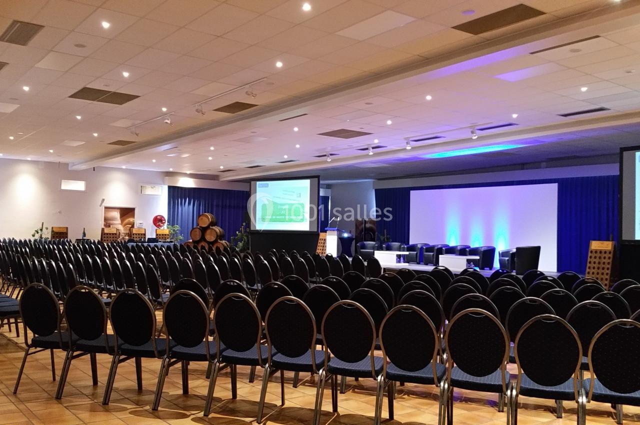 Salle de conférence aménagée avec des rangées de chaises noires, un podium et des écrans de projection allumés.