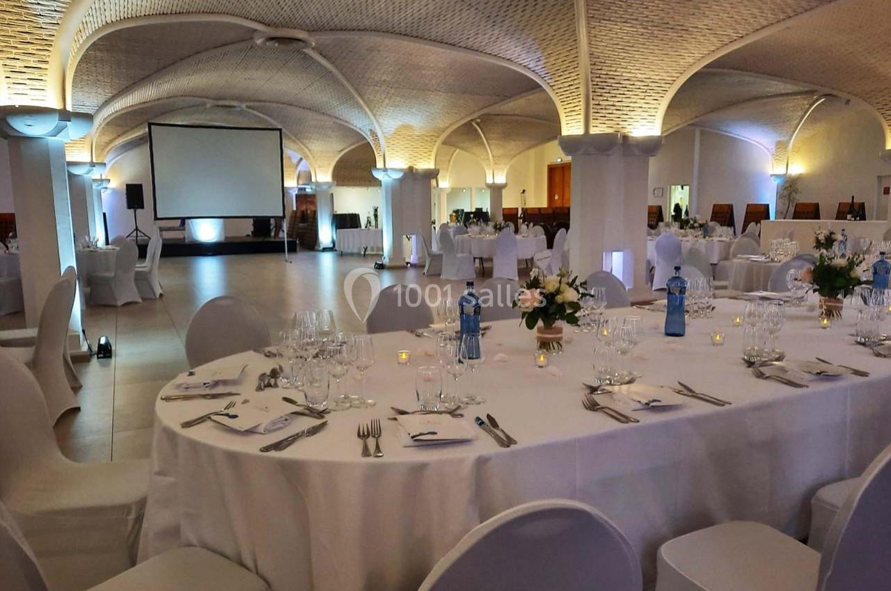 Salle de réception élégante avec tables rondes dressées, décor floral et éclairage tamisé sous un plafond voûté.