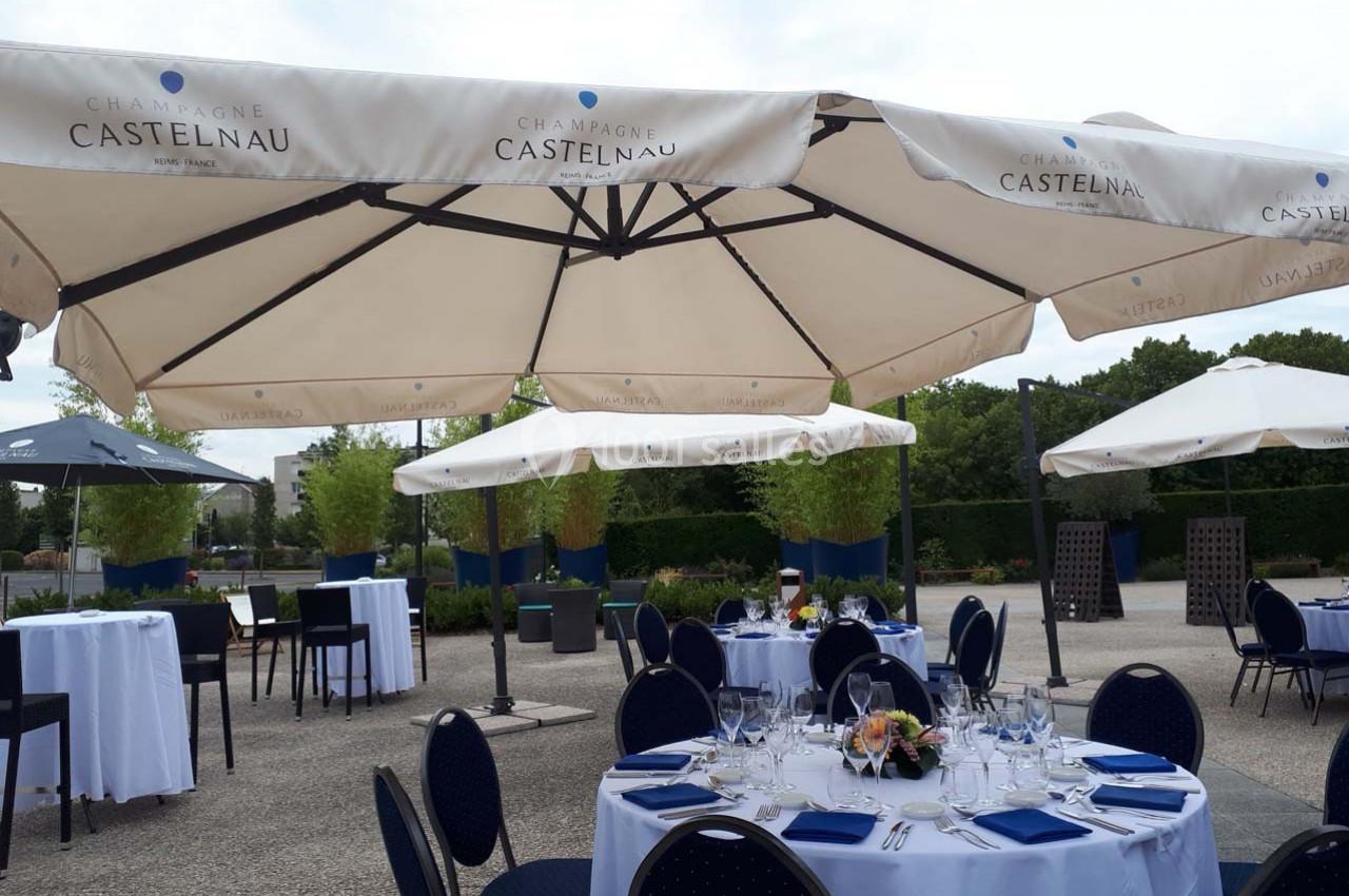Tables dressées avec nappes blanches et parasols blancs sur une terrasse extérieure, entourée de verdure.