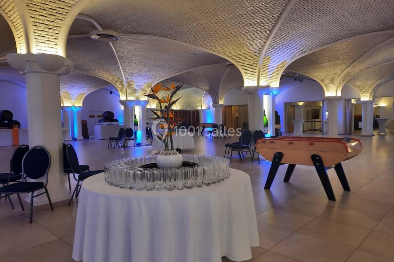 Salle voûtée avec tables dressées, chaises, baby-foot et éclairage d'ambiance coloré.