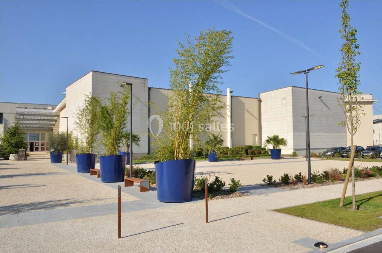 Bâtiment moderne entouré de grands pots bleus contenant des bambous, avec une allée pavée et un ciel dégagé.