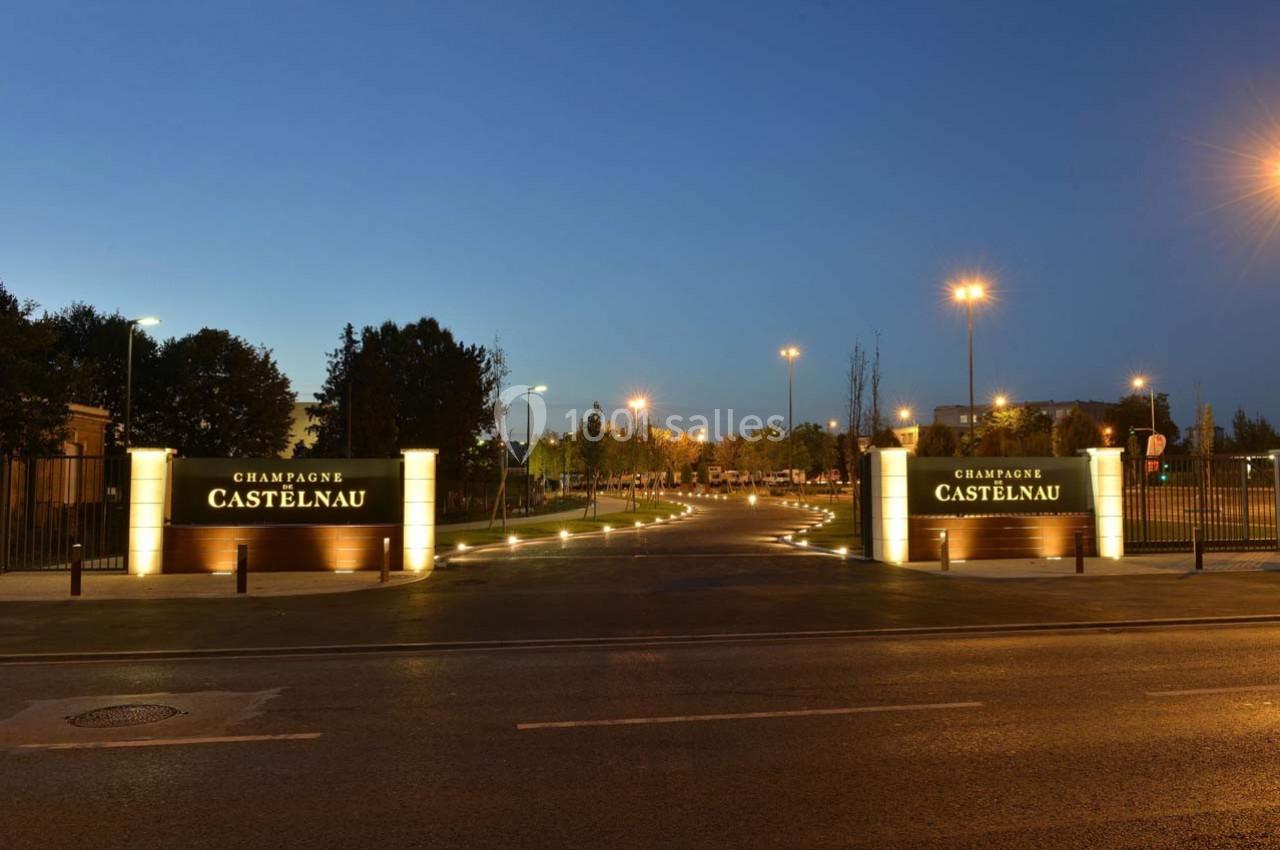Entrée éclairée du domaine Champagne Castelnau avec des panneaux lumineux et une route bordée d'arbres.