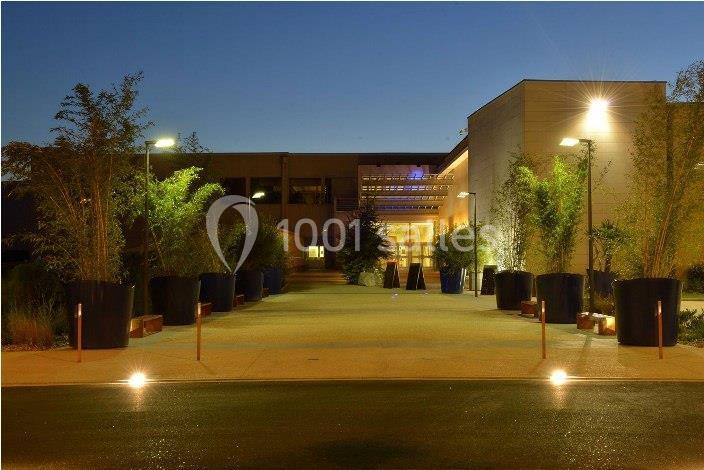 Entrée d'un bâtiment moderne éclairée de nuit, bordée de grands pots de plantes et d'allées pavées.