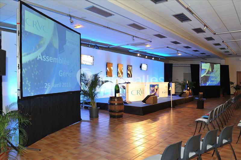 Salle de conférence avec scène éclairée, écrans projetant des informations, chaises alignées et décorations végétales.