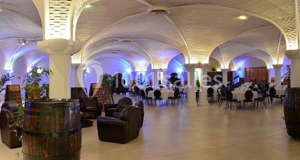 Salle voûtée éclairée avec des tables dressées, des chaises noires, des tonneaux en bois et des plantes décoratives.