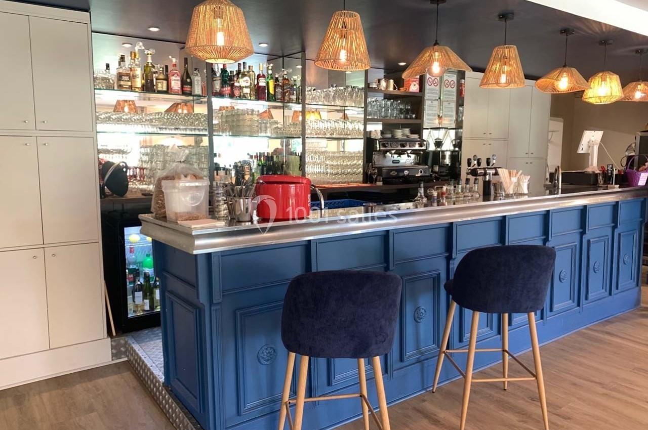 Comptoir de bar bleu avec tabourets, étagères de bouteilles, machine à café et suspensions lumineuses en rotin.