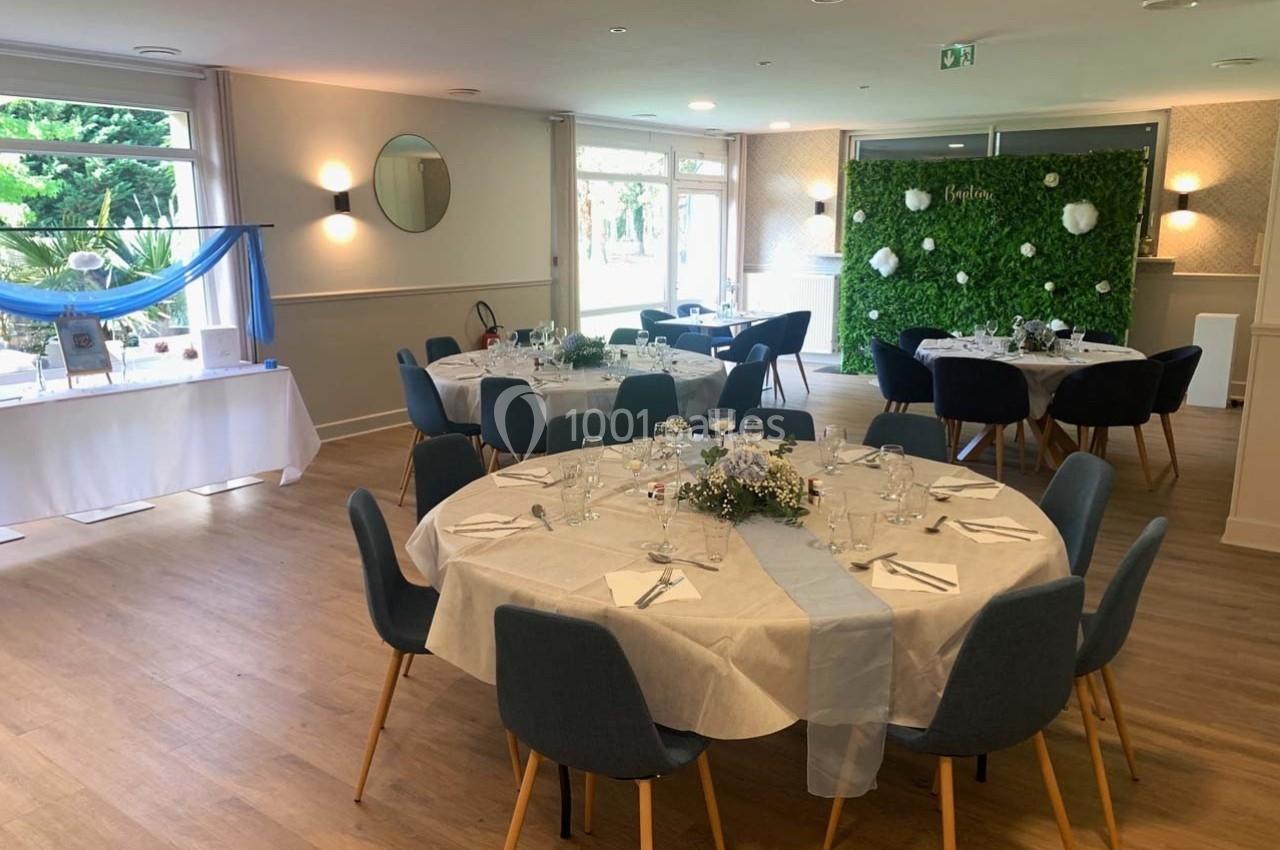 Salle de réception lumineuse avec tables rondes dressées, décorations florales et mur végétal en arrière-plan.