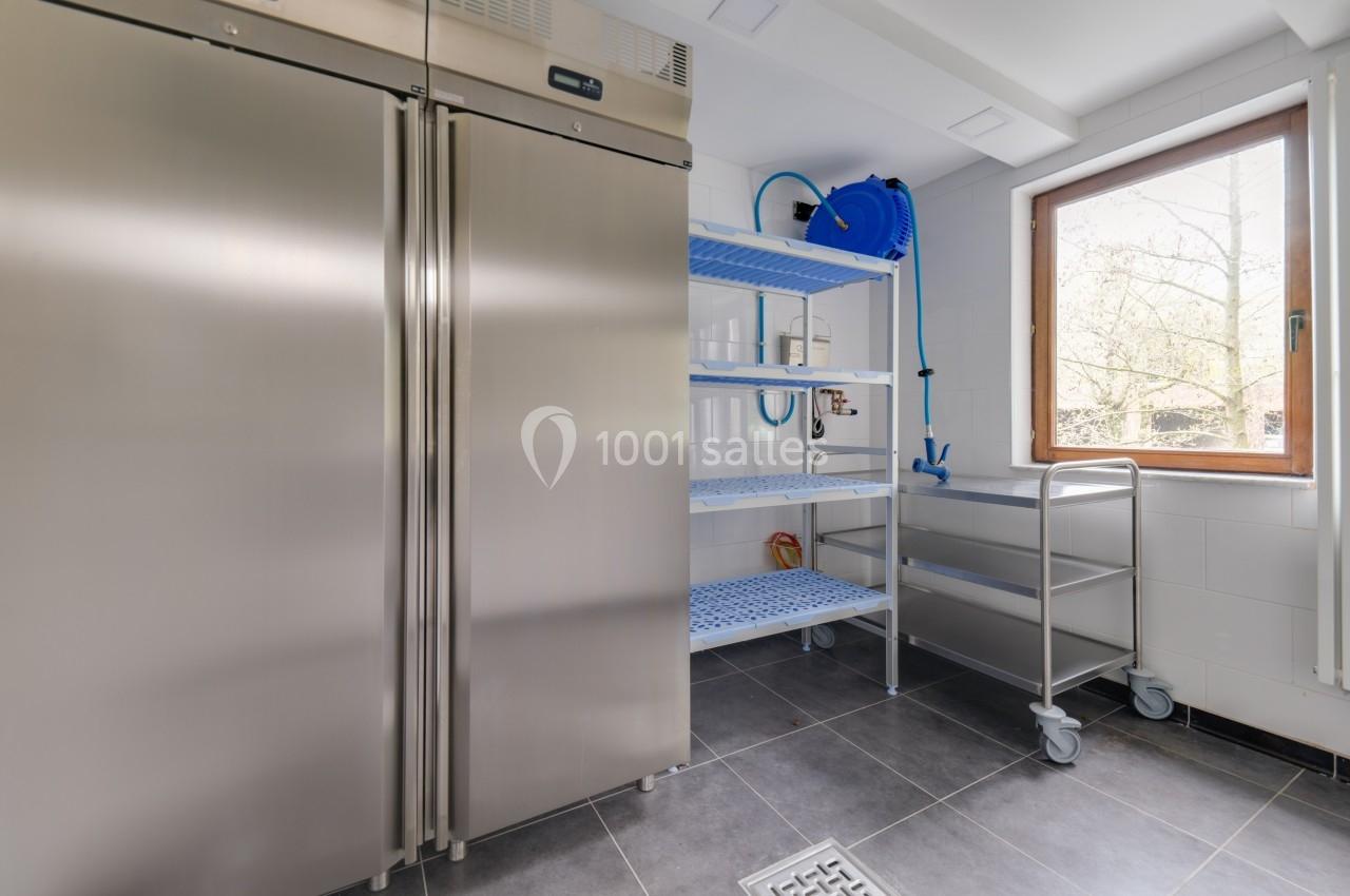 Salle de stockage avec réfrigérateur en inox, étagères métalliques bleues et chariot sur roulettes près d'une fenêtre.