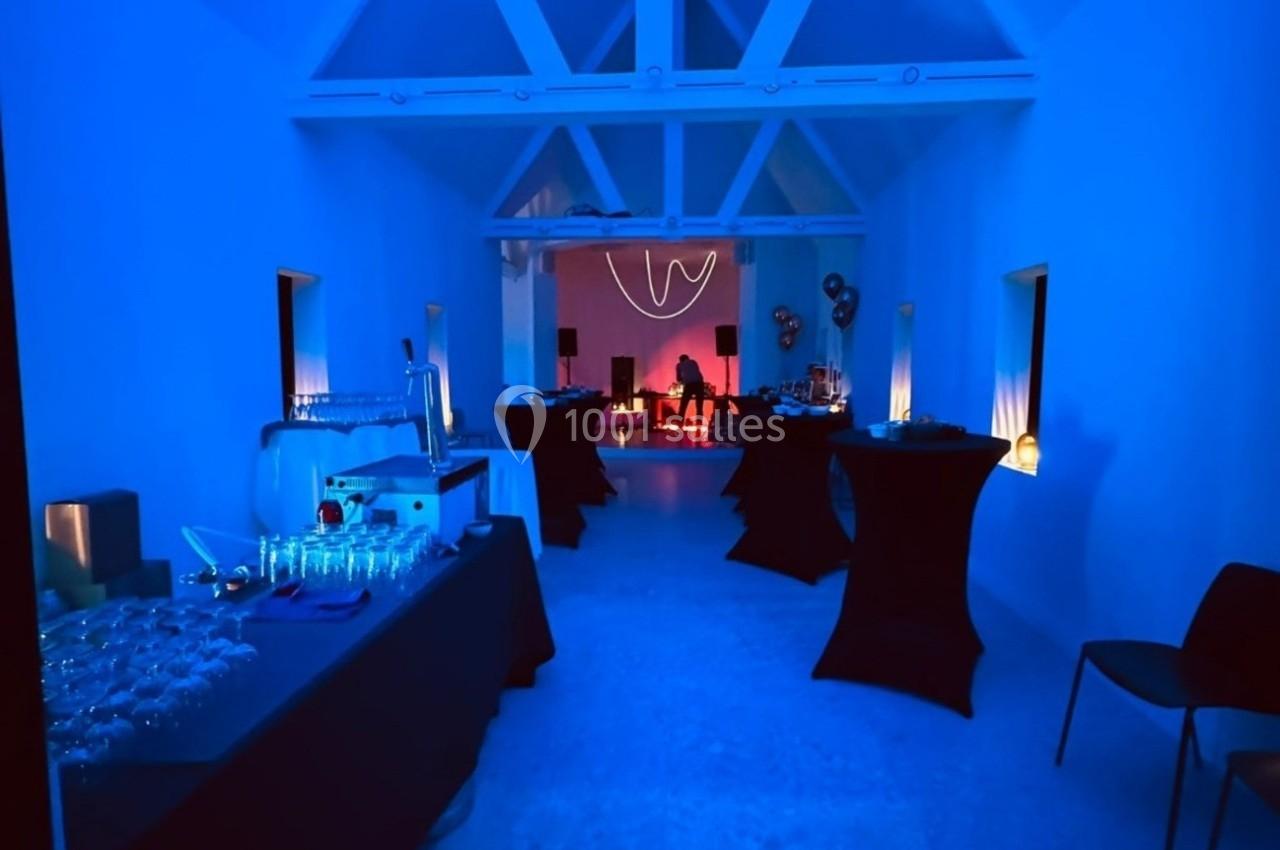 Salle éclairée en bleu avec tables dressées pour un événement, et une zone rouge illuminée en arrière-plan.