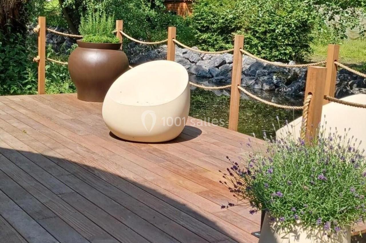 Terrasse en bois avec fauteuils modernes, pots de fleurs et vue sur un jardin verdoyant près d'un cours d'eau.