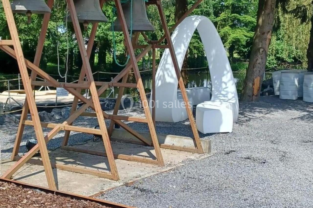 Structure en bois avec des cloches suspendues, entourée d'arbres et d'une arche blanche dans un espace extérieur.