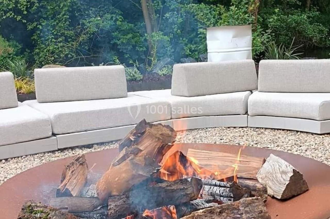 Brasero en métal avec des bûches enflammées, entouré de fauteuils beige sur un sol en gravier, dans un jardin boisé.