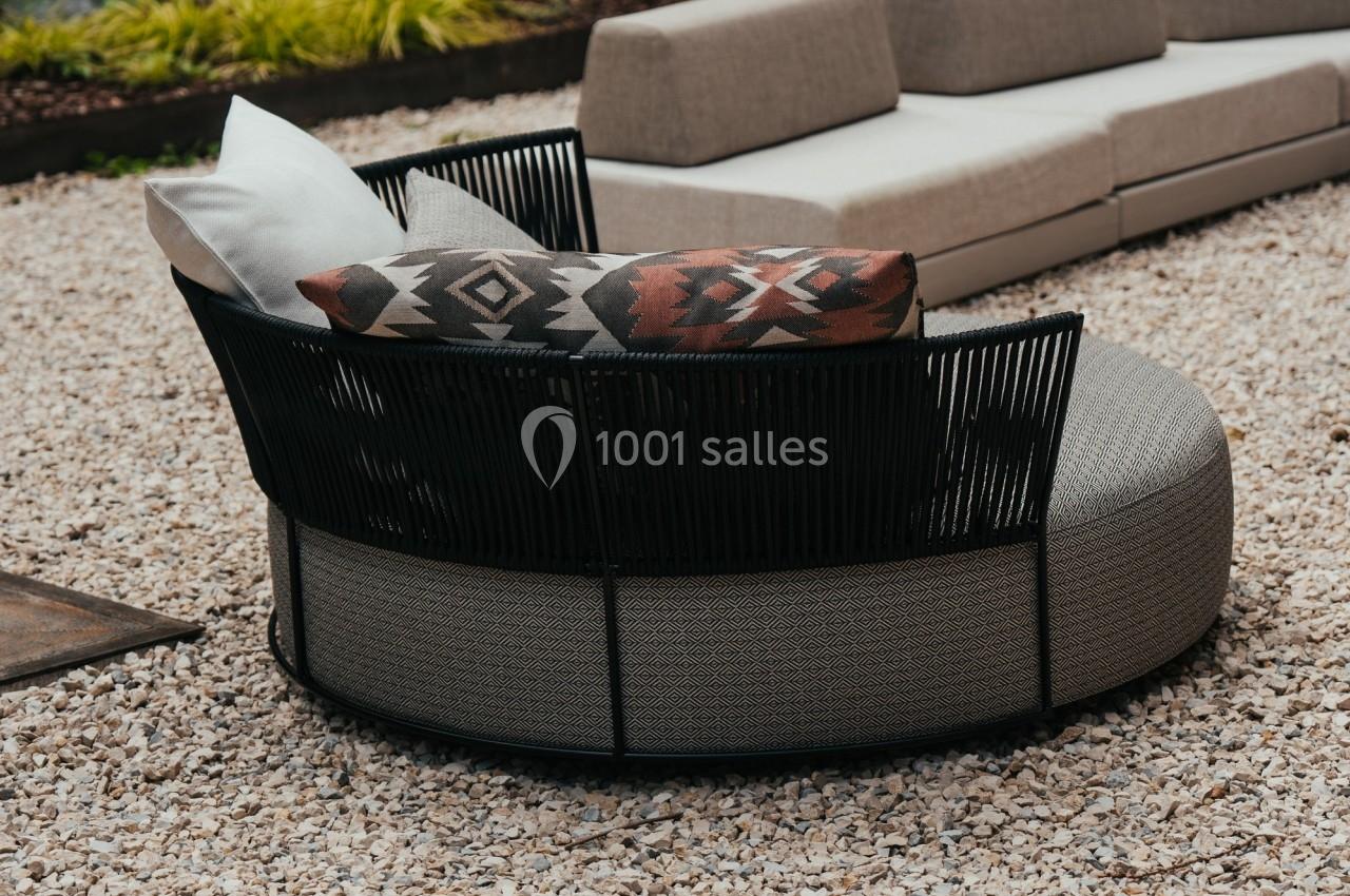 Fauteuil rond en rotin noir avec coussins, placé sur un sol en gravier dans un jardin avec canapé en arrière-plan.