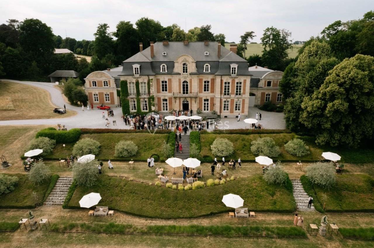 Vue aérienne d'un château entouré de jardins, avec des invités rassemblés devant l'entrée principale.