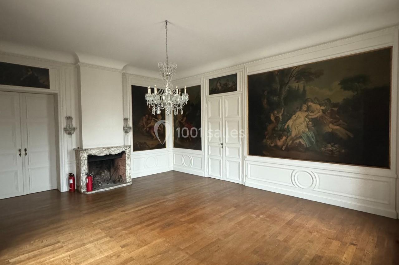Salle vide avec parquet en bois, murs ornés de grandes peintures, cheminée en marbre et lustre suspendu.