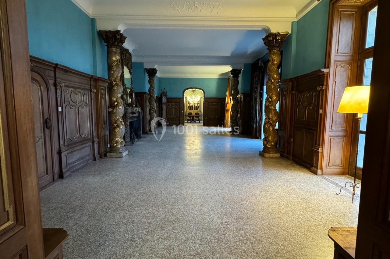 Vue d'un couloir spacieux avec murs bleus, colonnes décoratives, parquet en mosaïque et lumière naturelle entrant par les…