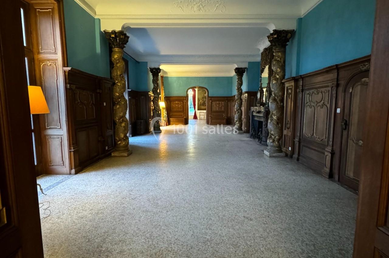 Vue d'un couloir spacieux avec murs bleus, colonnes décoratives, parquet clair et portes en bois sculpté.