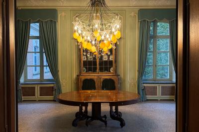 Salle élégante avec un lustre imposant, table ronde en bois, chaises vertes et grandes fenêtres encadrées de rideaux.