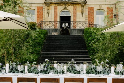 Une mariée en robe longue marche aux côtés d'un homme en costume sur une terrasse avec vue sur un paysage verdoyant.