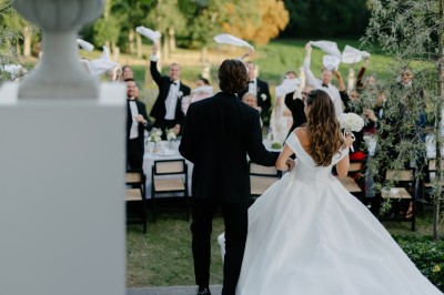 Une mariée en robe longue marche aux côtés d'un homme en costume sur une terrasse avec vue sur un paysage verdoyant.
