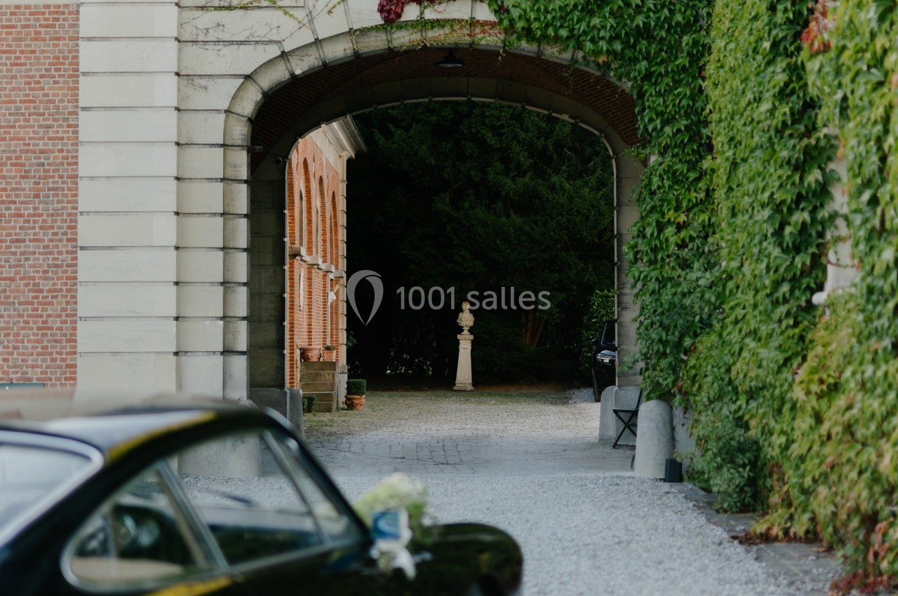 Voûte en pierre entourée de murs recouverts de lierre, donnant sur une allée pavée avec une statue au centre.