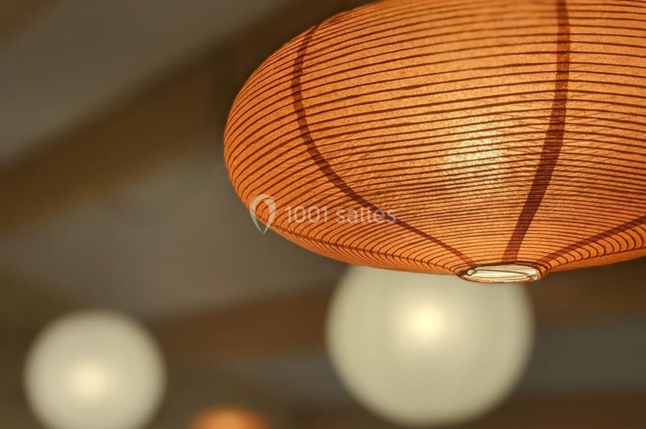 Suspension lumineuse en papier orange avec des lampes floues en arrière-plan dans une ambiance chaleureuse.