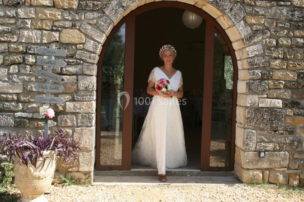 Femme en robe de mariée tenant un bouquet, debout devant une porte voûtée en pierre.
