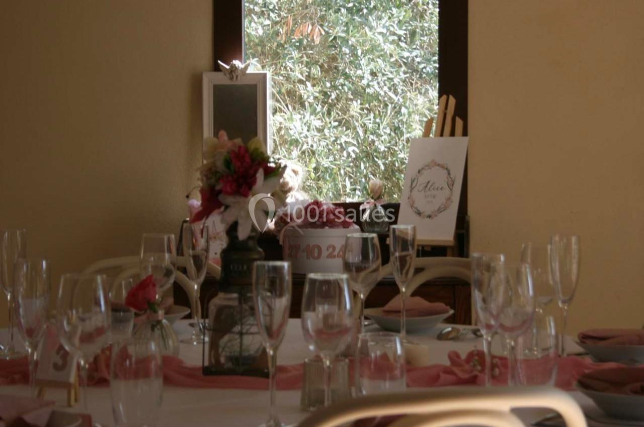 Table décorée pour un événement avec nappes blanches, verres, fleurs roses et fenêtre donnant sur des arbres.