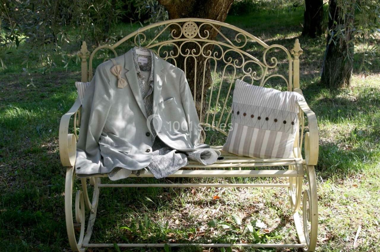 Veste grise et coussin rayé posés sur un banc en métal beige dans un jardin ombragé.