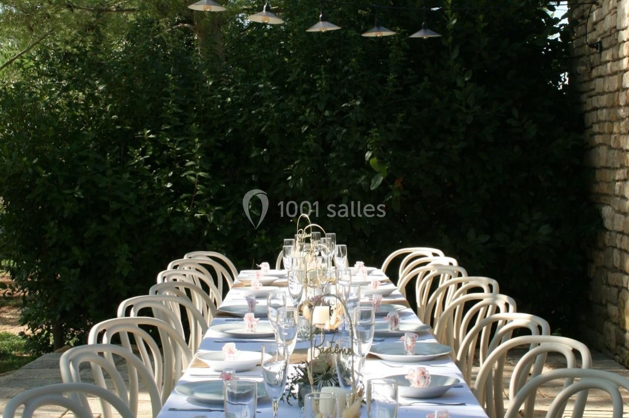 Table extérieure dressée pour un repas, avec nappes blanches, chaises assorties et décoration élégante sous des lampes…