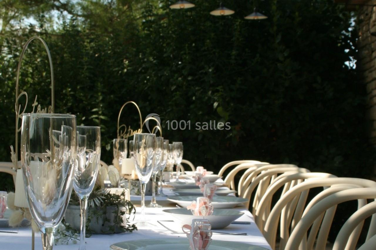 Table dressée en extérieur avec vaisselle élégante, verres et décorations florales, entourée de chaises blanches.