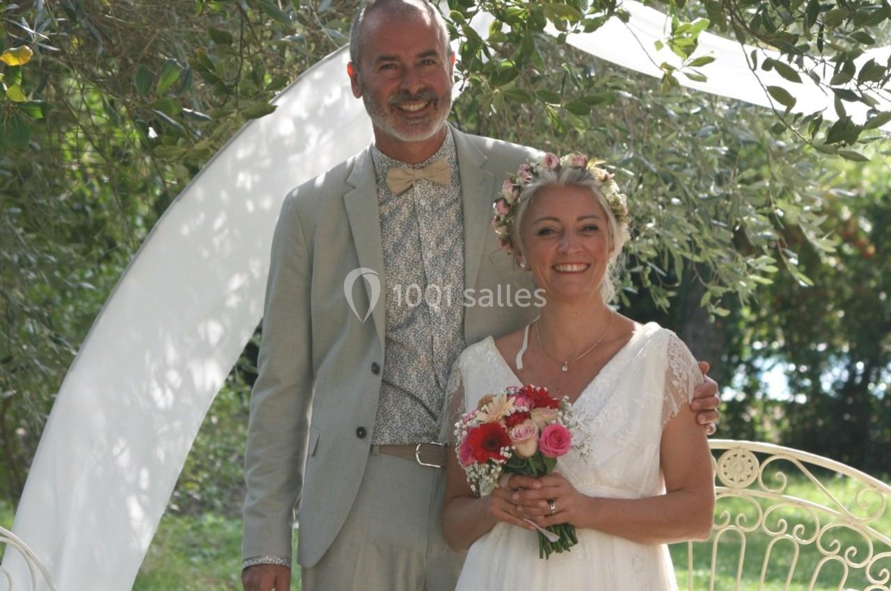 Un couple souriant en tenue de mariage pose dans un jardin, entouré de verdure et d'une arche blanche.